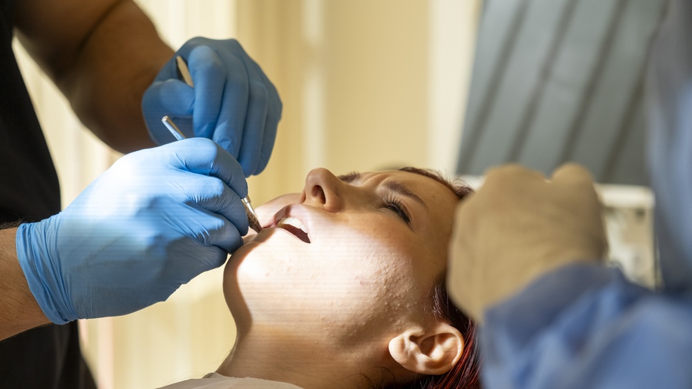 A person lies in a dental chair with their mouth open while two gloved dentists work, using dental instruments to examine or treat their teeth in a brightly lit clinic.