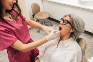 A dentist in pink scrubs examines the open mouth of an older woman with gray hair and glasses, who sits in a dental chair. The dentist wears gloves, and dental tools are visible in the clean clinic setting.